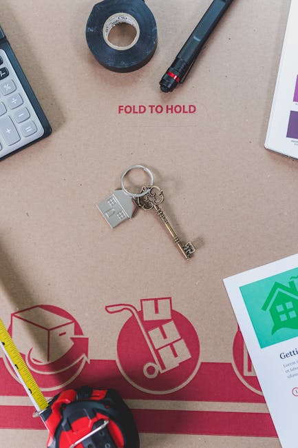 A close-up view of a brown cardboard moving box with various objects placed on top, including a roll of dark packaging tape, a black marker pen, and a set of house keys with a small silver house-shape