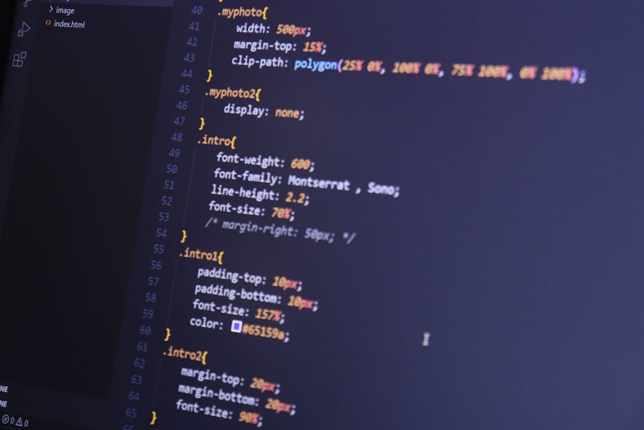 A close-up image showing a person's right hand using a laptop on a wooden surface, with the screen displaying lines of code. The individual is wearing a wristwatch with a patterned strap on their left