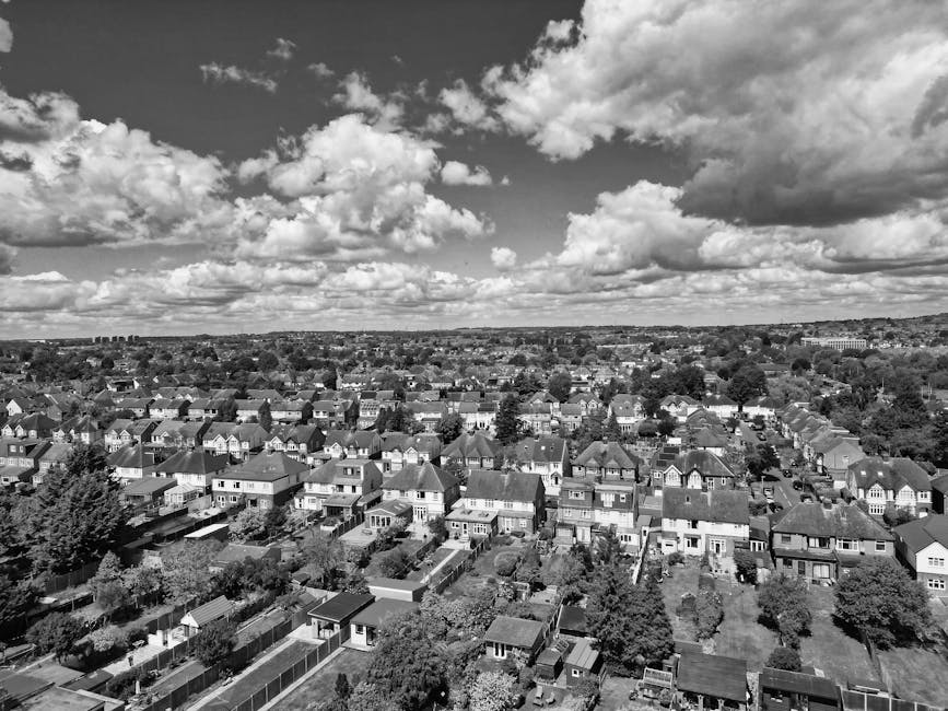 Black and white aerial photograph showing a residential area during a home relocation process. In the foreground, several cardboard boxes and wrapped pieces of furniture are positioned on a driveway a