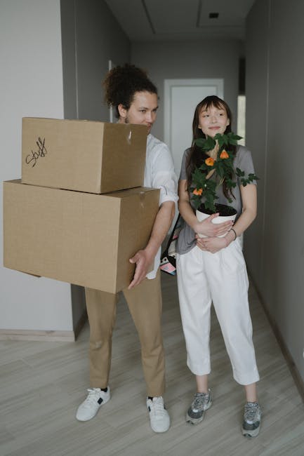A person wearing white trousers and an orange top is inside a room, stacking and arranging large cardboard boxes on a concrete floor against a plain white wall. Several boxes of varying sizes are posi